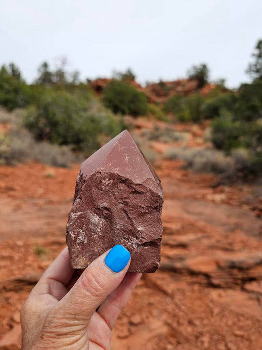 A hand holding a cut base point of Red Jasper with a desert landscape in the background.